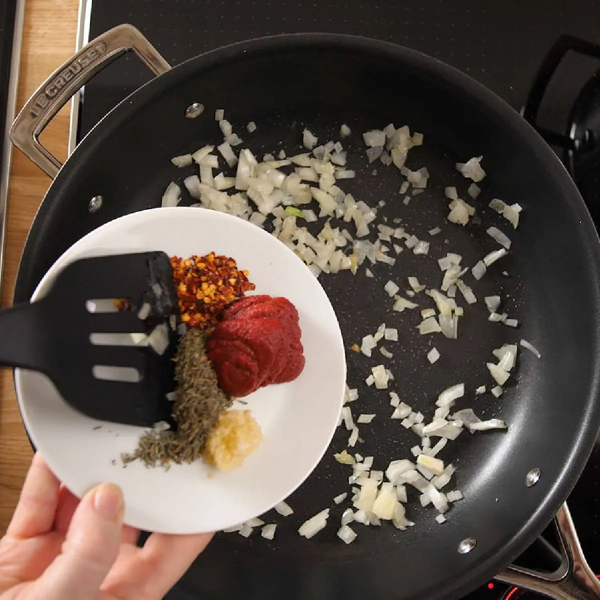 A hand uses a black spatula to add spices (chili flakes, dried herbs, garlic paste) and tomato paste from a white plate into a frying pan with sautéed onions.