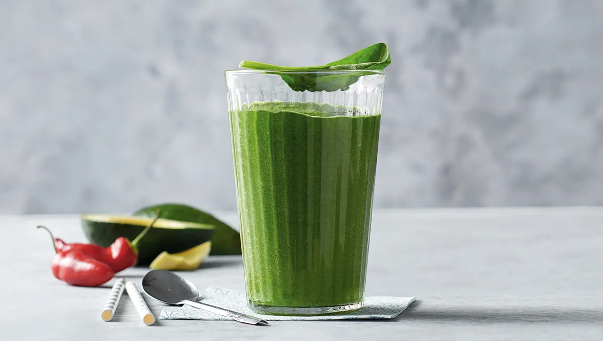 Frullato verde in un bicchiere disposto insieme a metà avocado e un peperoncino.