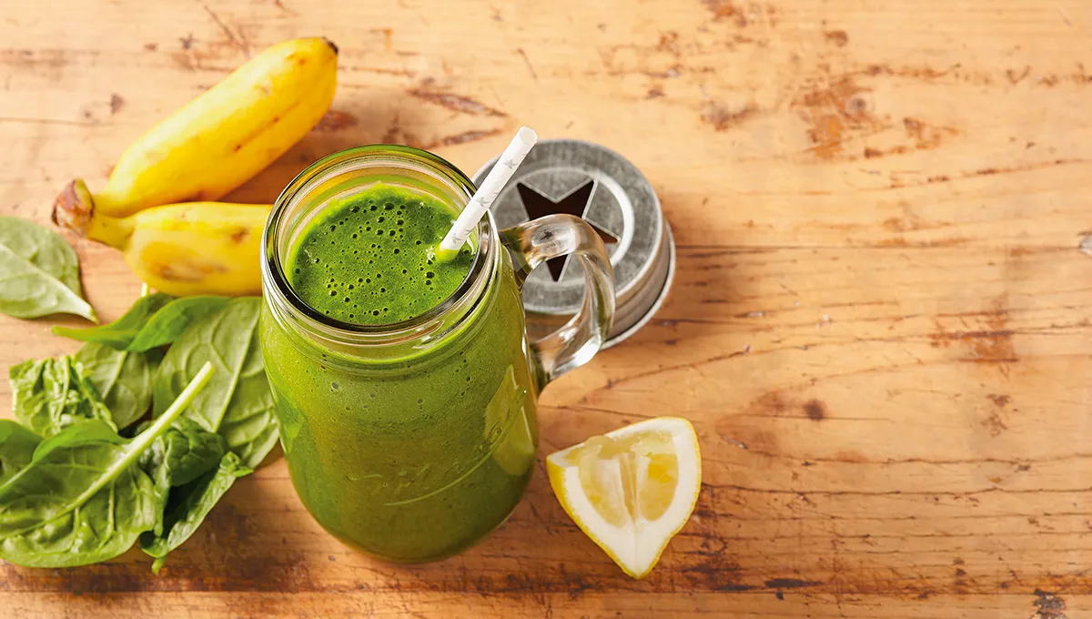Frullato verde in un bicchiere disposto insieme a foglie di spinaci, banane e una fetta di limone su un tavolo.