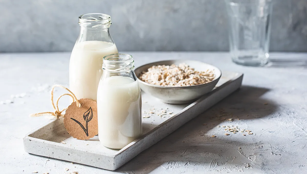 Due bottiglie di vetro di latte d'avena su un vassoio con una ciotola di avena sullo sfondo, adagiate su una superficie dalla consistenza leggera.