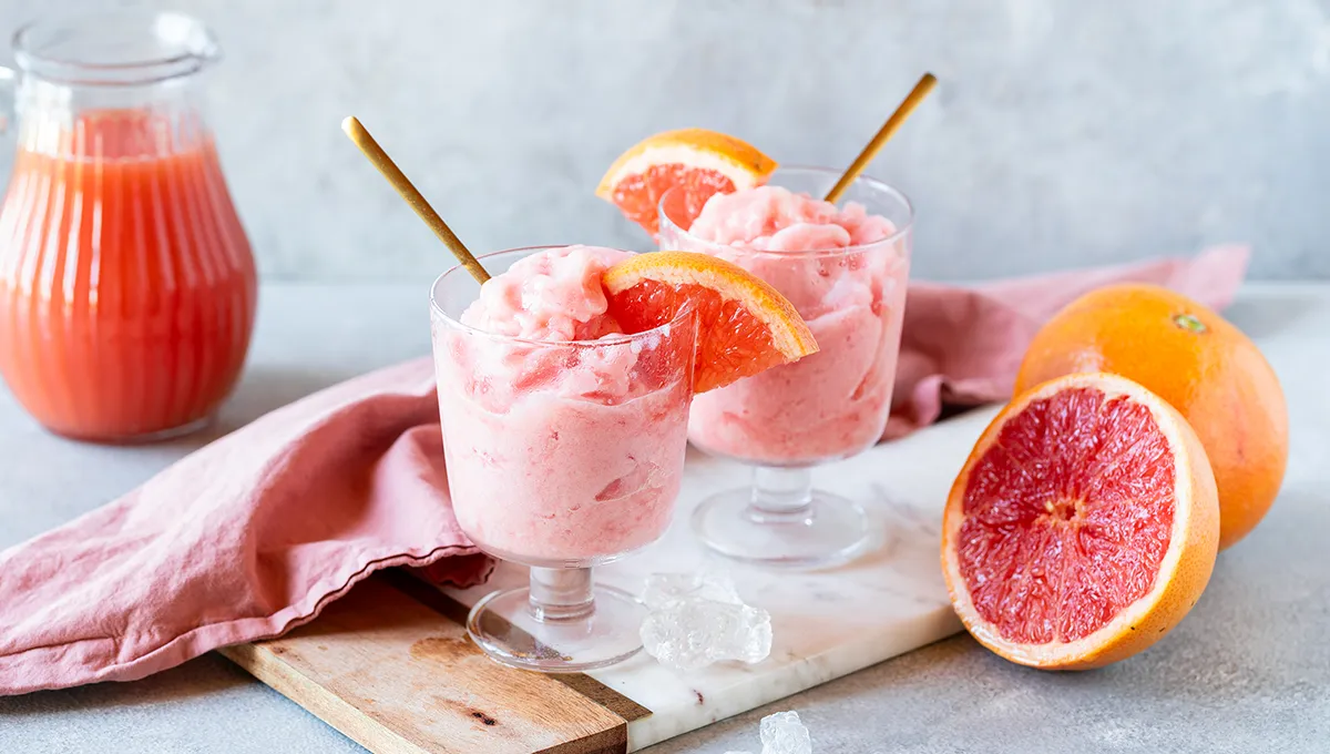 Due bicchieri di cremosa granita al pompelmo su un tavolo di legno scuro, ciascuno guarnito con una fetta di pompelmo fresco.
