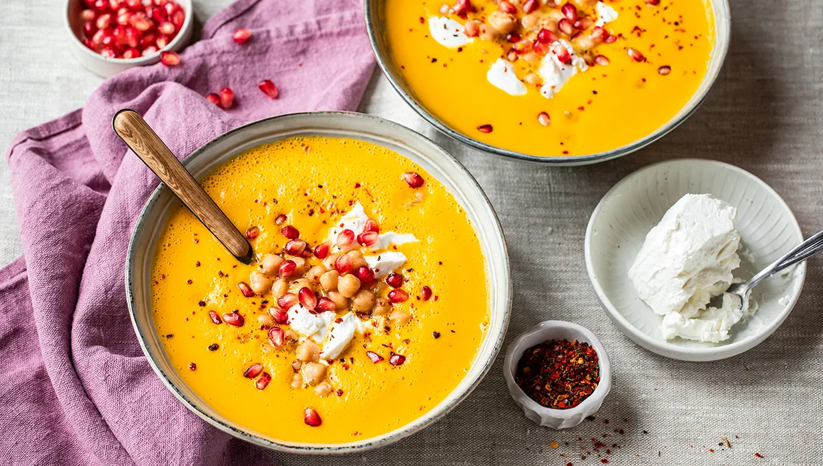 Ciotole di zuppa di patate dolci condite con ceci, semi di melograno e un ciuffo di panna, disposte su un tavolo con un tovagliolo viola e un contorno di panna e semi di melograno.