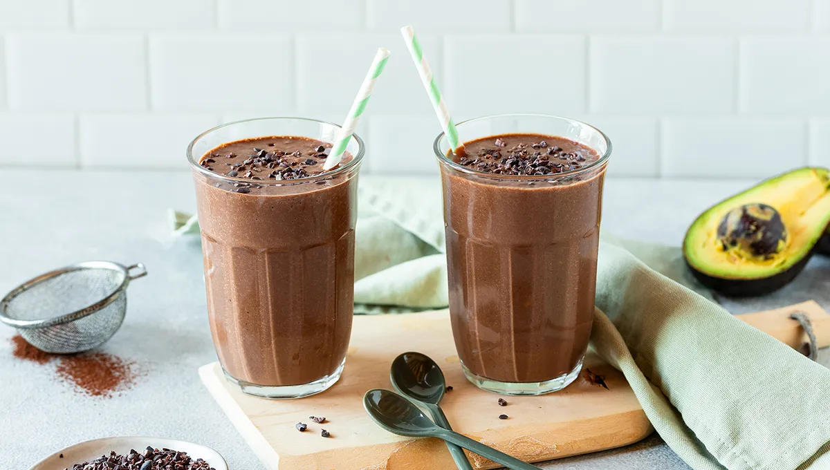 Due bicchieri di frullato di cioccolato e avocado con fave di cacao su un vassoio scuro con un cucchiaio di fave accanto.