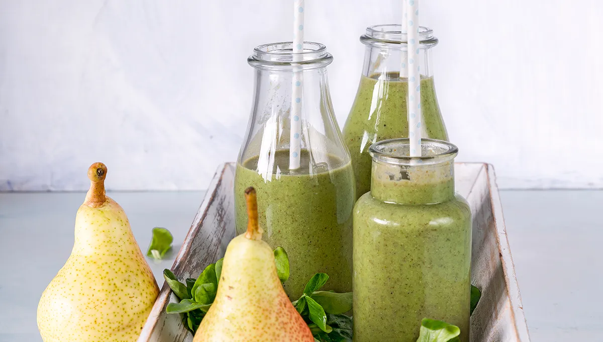 Quattro bottiglie di frullato verde, ciascuna con una cannuccia, vengono poste in un vassoio bianco con due pere e foglie verdi.