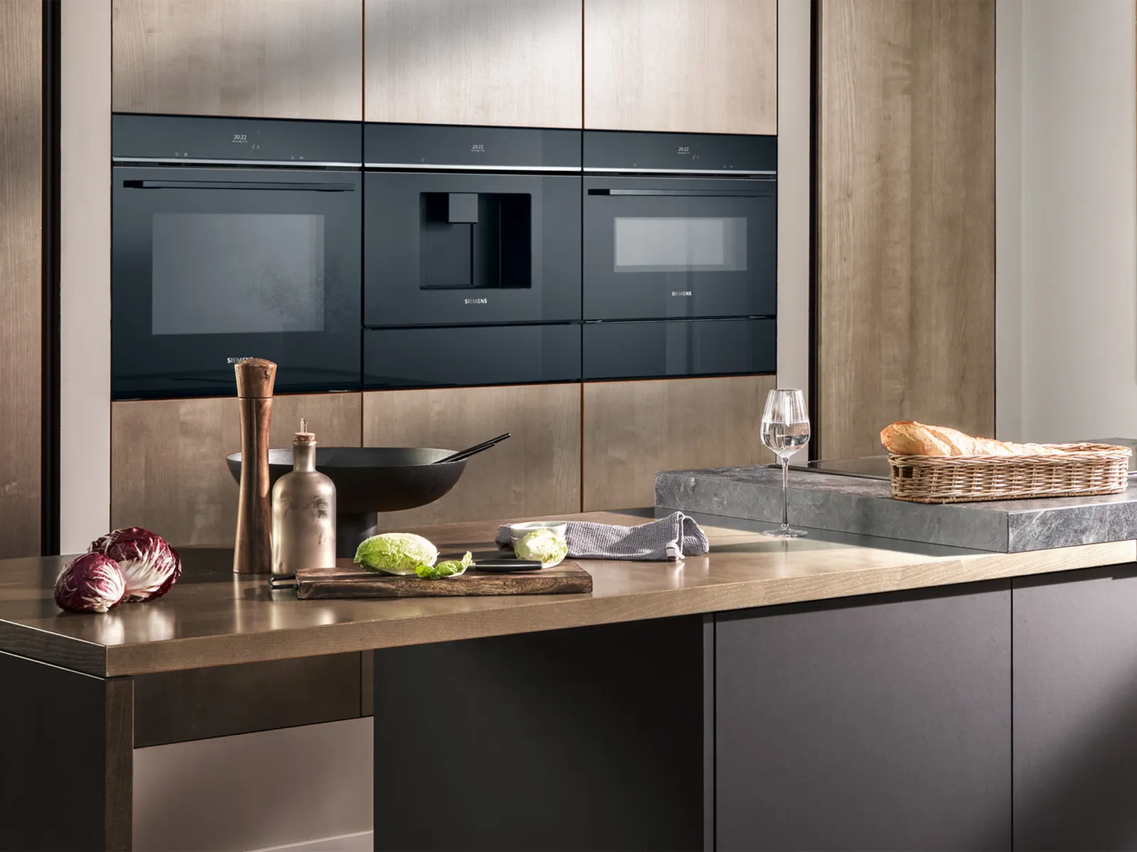 Interior de cozinha moderna com eletrodomésticos Siemens encastrados, bancada de madeira, legumes, cesta de pão, copo de vinho e utensílios de cozinha.