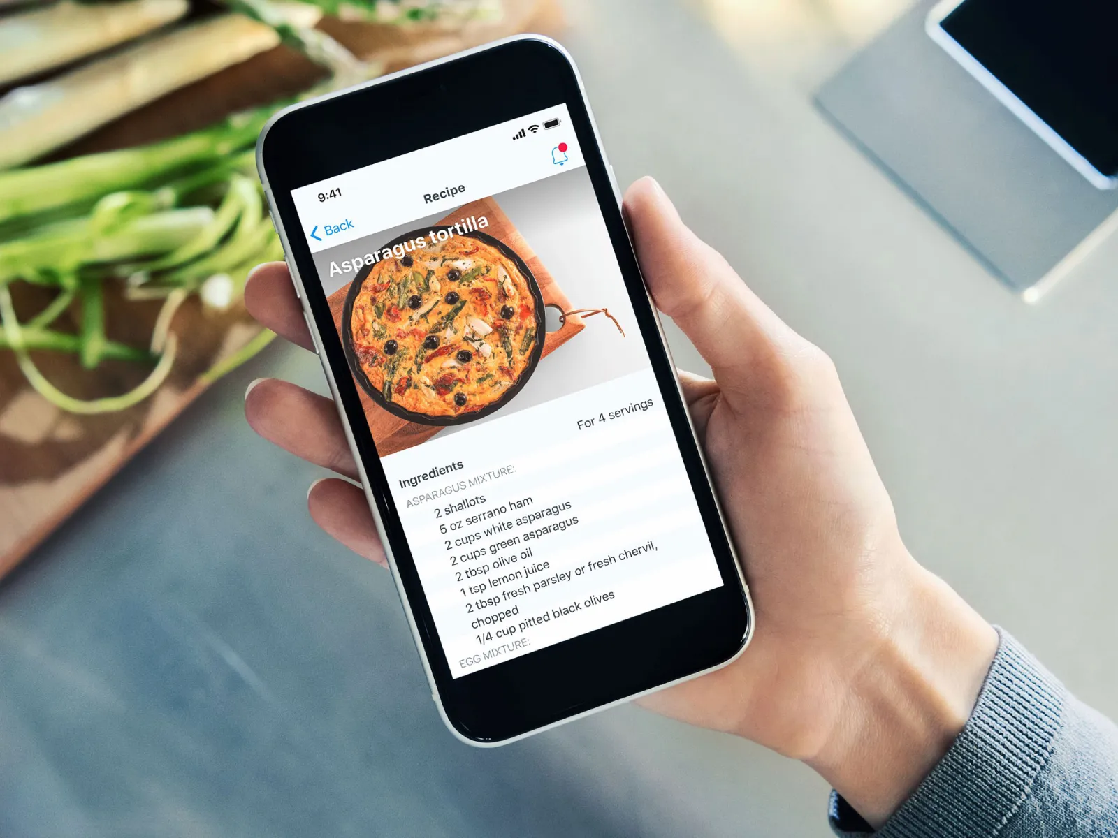 Immagine di una mano che impugna uno smartphone con una ricetta aperta sull‘app Siemens Home Appliances; sullo sfondo, alcuni asparagi freschi su un tagliere in legno.