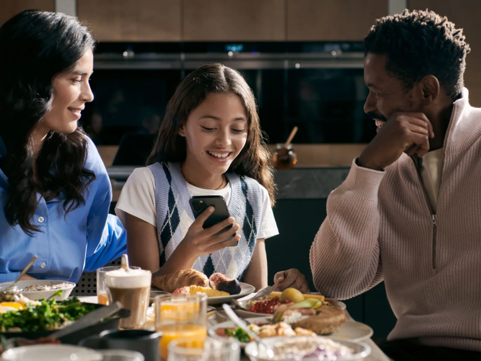 Três pessoas a desfrutar de uma refeição em conjunto, uma a usar um smartphone e as outras a conversar.