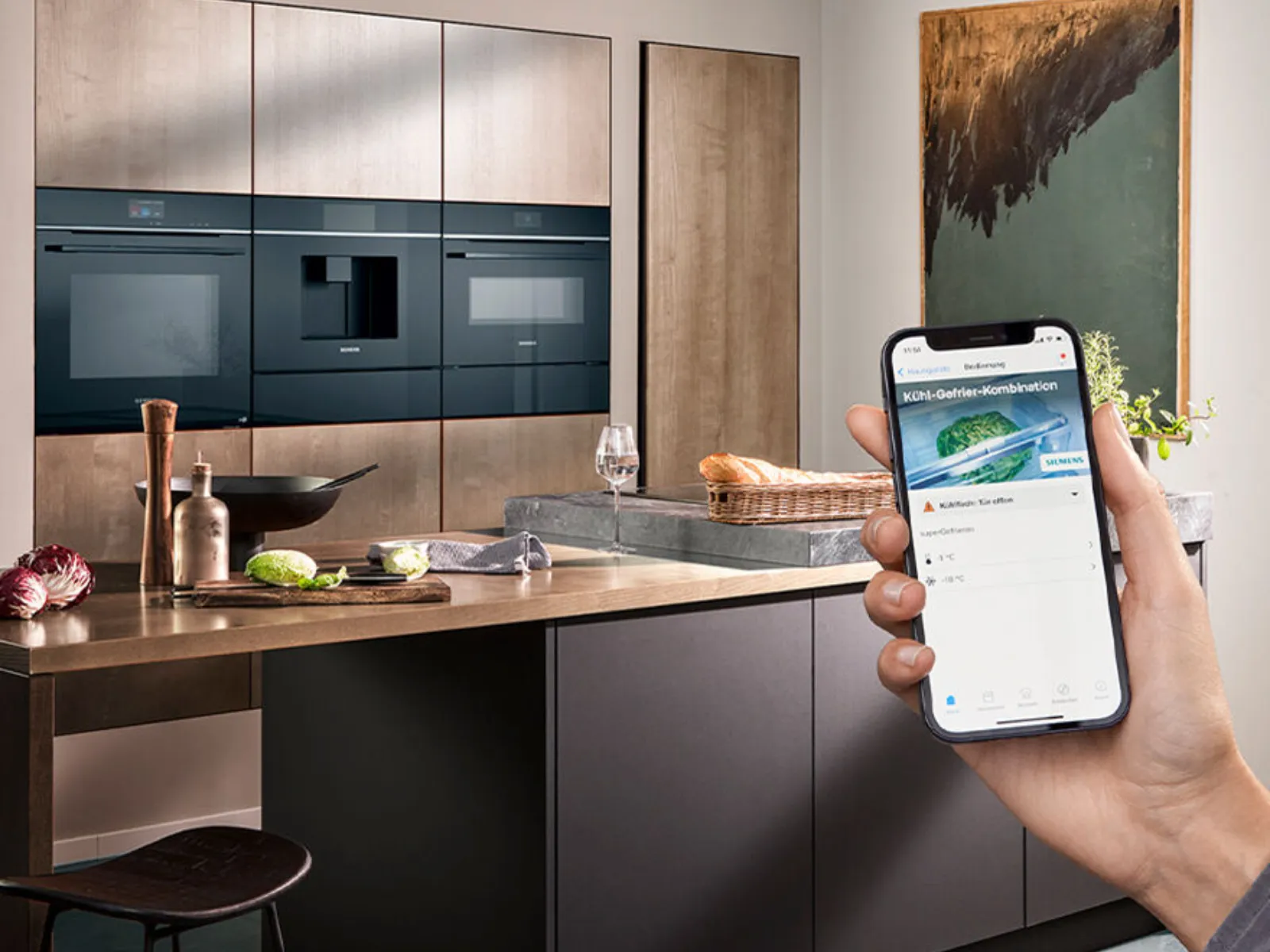 Modern kitchen with built-in Siemens oven, coffee machine, microwave. Hand holds phone showing Siemens fridge app with "door open" warning.