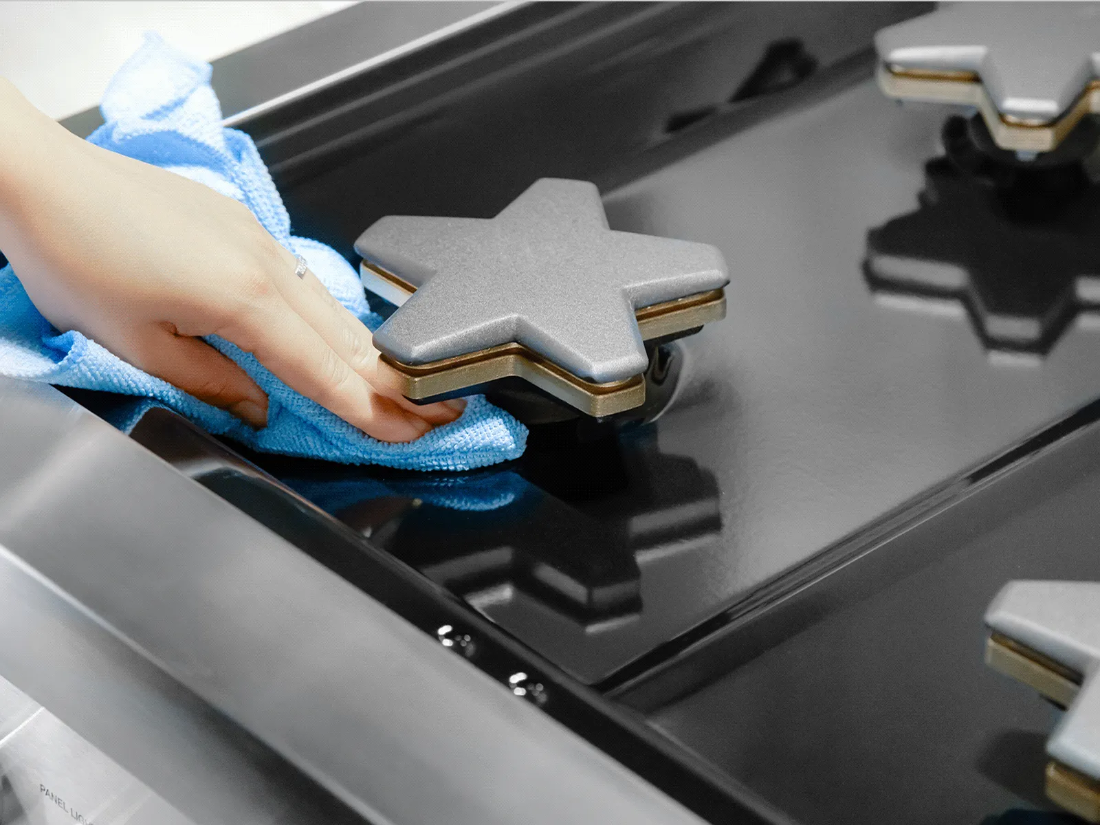 Hand wiping a glossy black gas stovetop with a blue cloth, showing star-shaped burner caps.