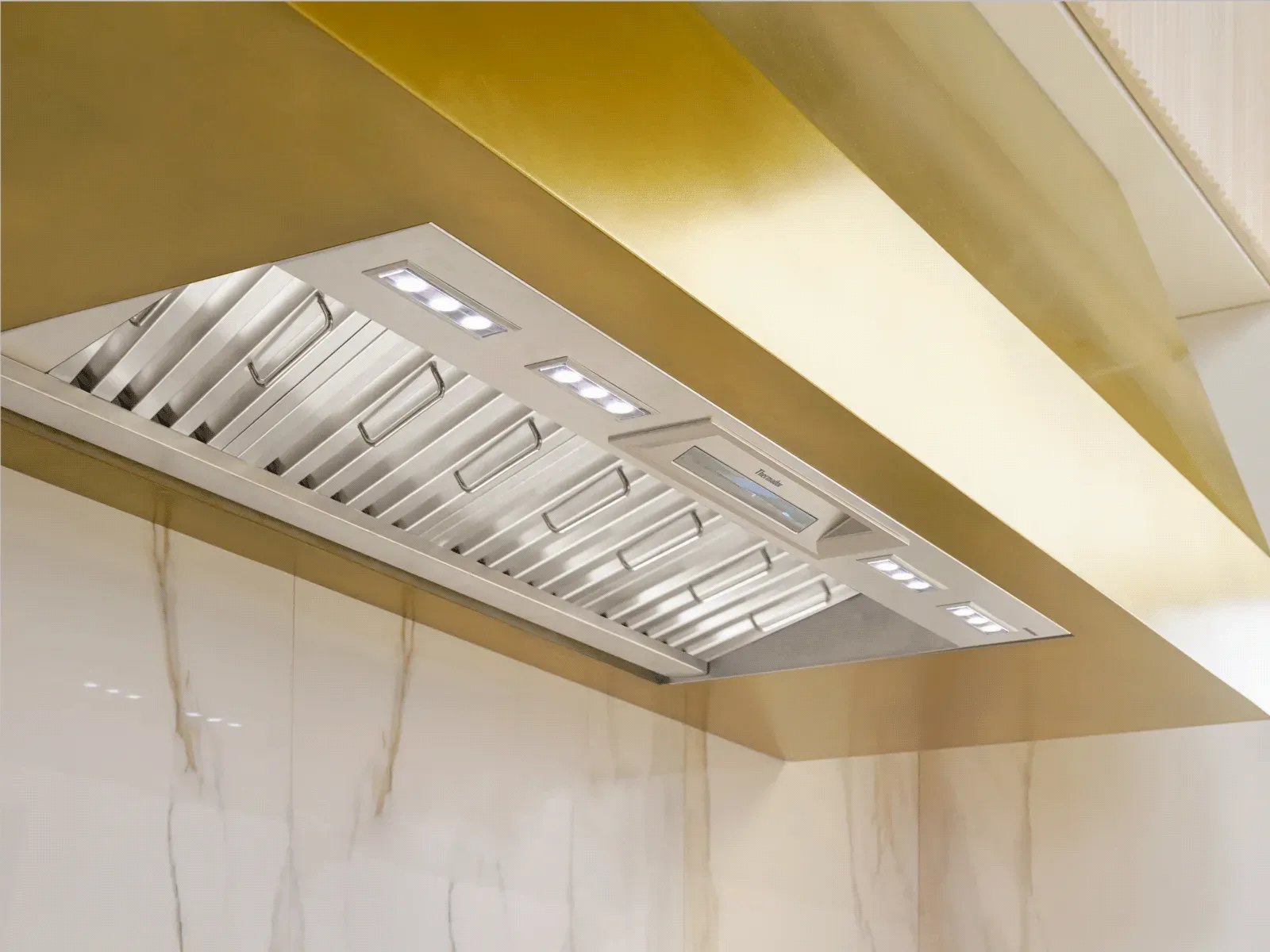 Under-cabinet stainless-steel range hood with baffle filters and built-in LED lights inside a gold-finish canopy over a marble backsplash.