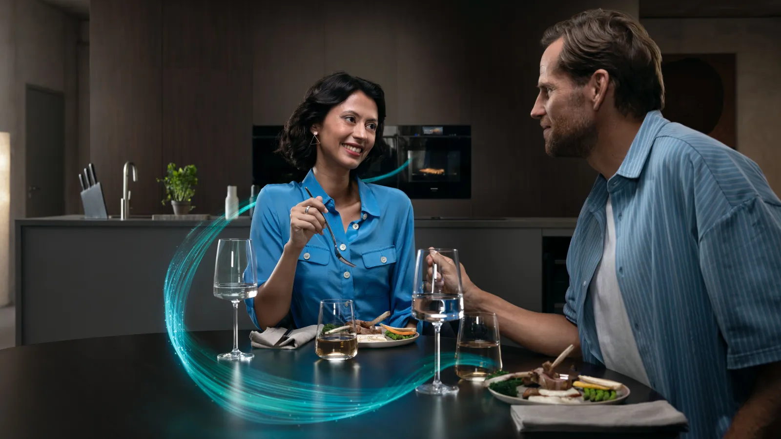 Due persone cenano insieme a un tavolo con piatti di cibo e bicchieri d'acqua, all'interno di una cucina moderna.