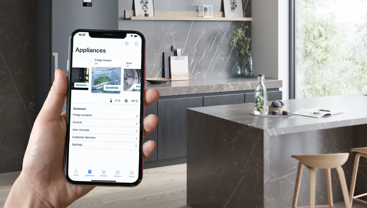 Hand holding smartphone displaying smart home app controlling Siemens fridge freezer and dryer in a modern kitchen setting.