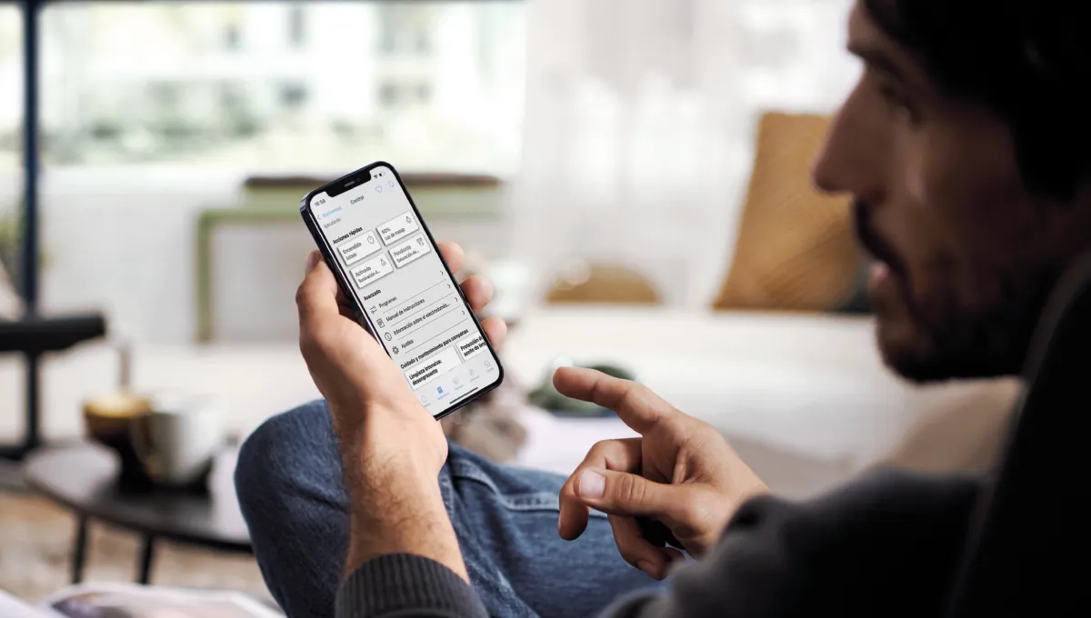 Man sitting and holding a smartphone displaying an application screen with control and settings options in Spanish.