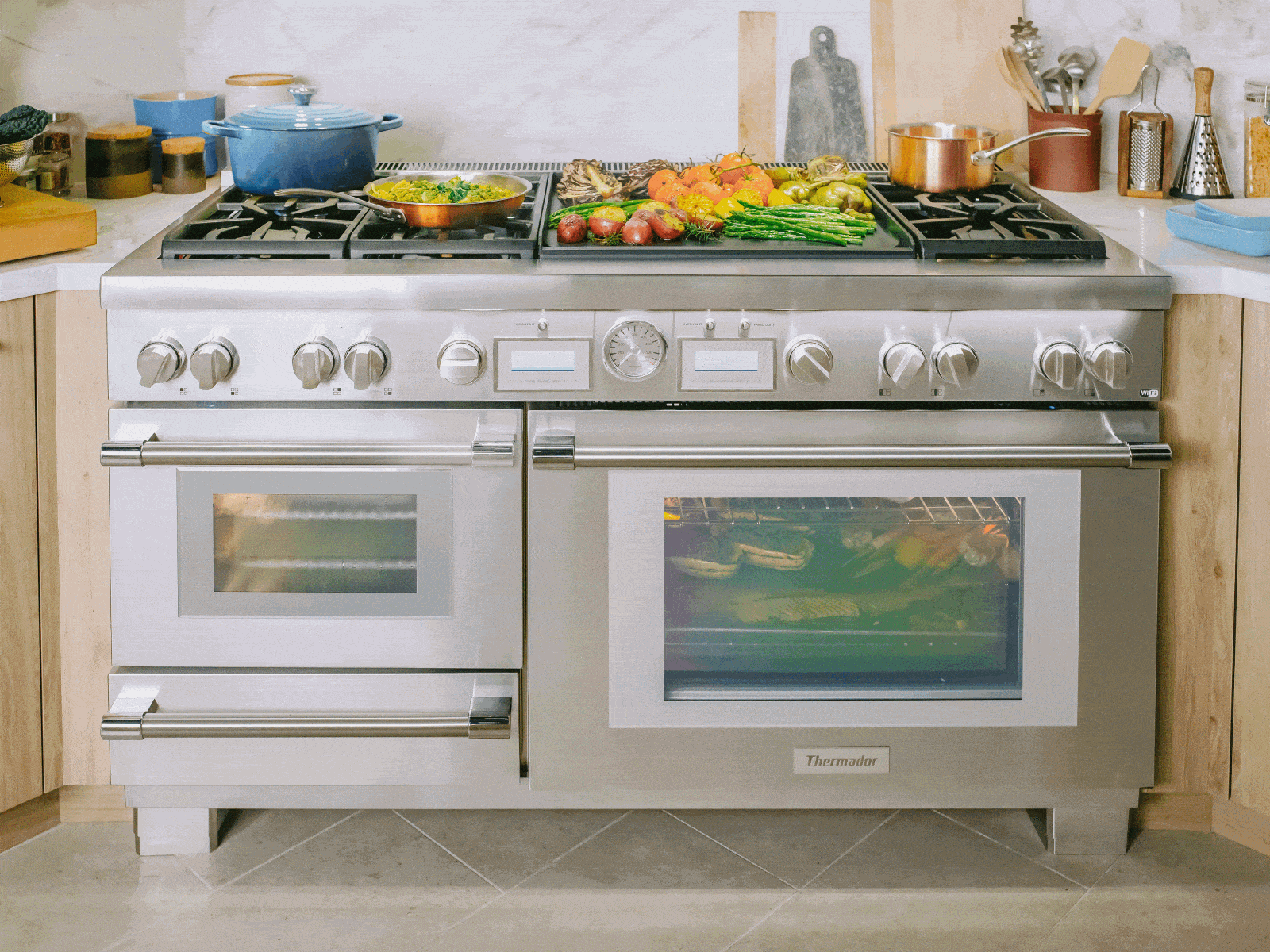 A large, stainless steel Thermador professional-style range is the centerpiece of a modern kitchen. Its versatile cooktop, featuring both gas burners and a griddle, is laden with colorful dishes like roasted vegetables and a blue Dutch oven. The appliance also includes a main oven, a steam oven, and a warming drawer, with food visible cooking through the main oven door.