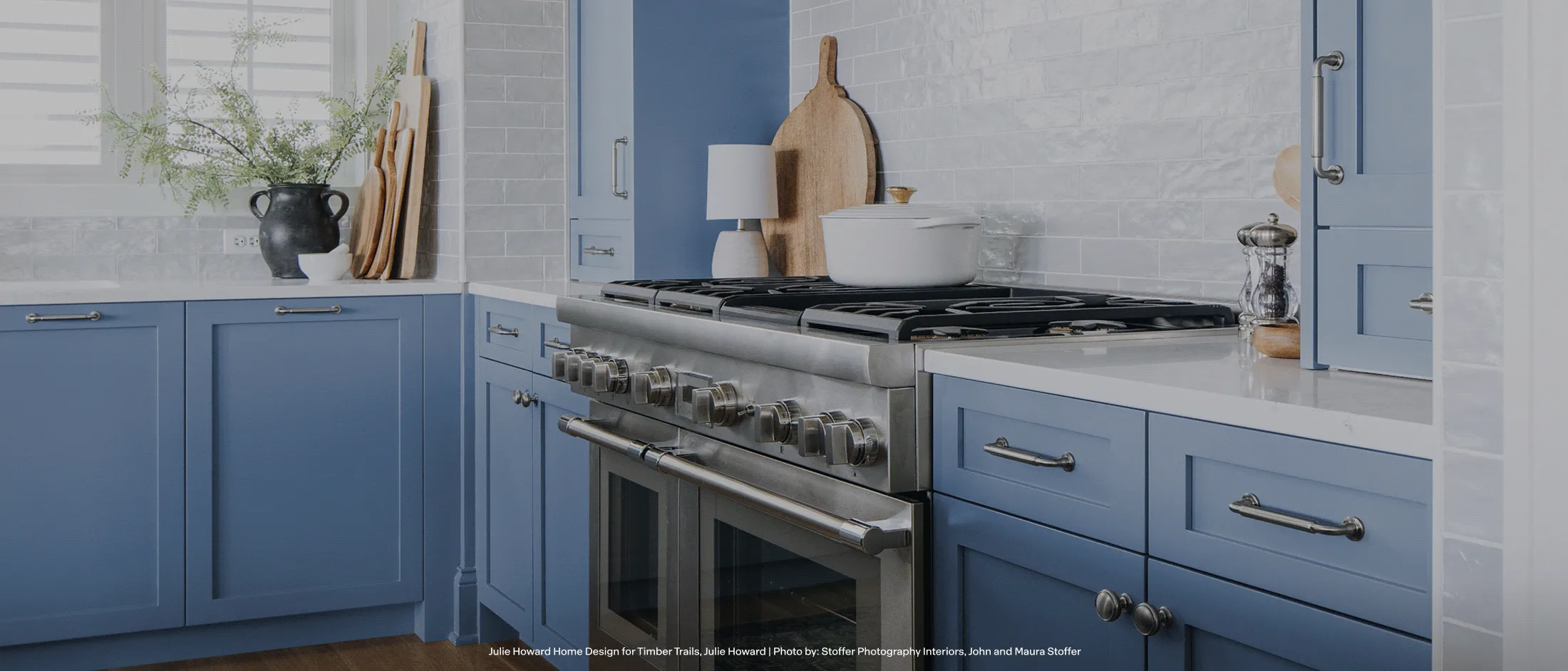 Modern kitchen with blue cabinets, stainless steel stove, white countertops, wooden cutting boards, and decorative black vase with greenery.