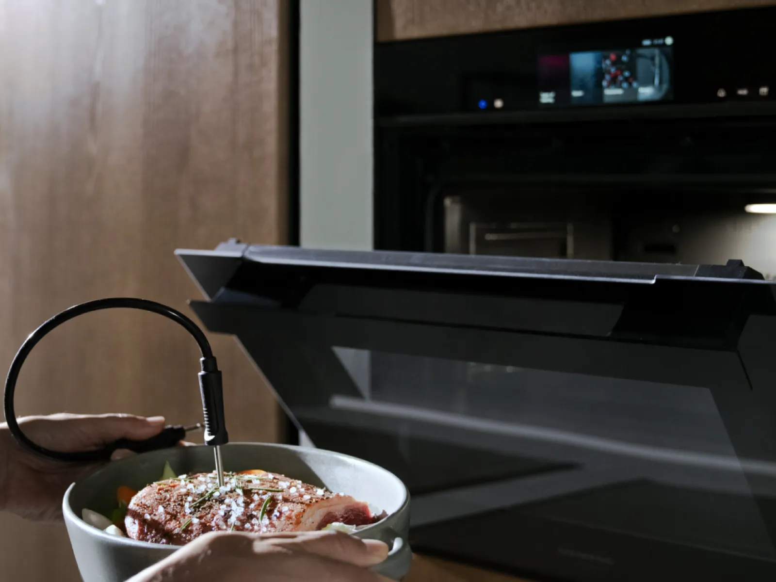 Des mains tenant un récipient contenant de la viande assaisonnée avec une thermosonde, prête à être enfournée dans un four ouvert doté d'un affichage numérique.