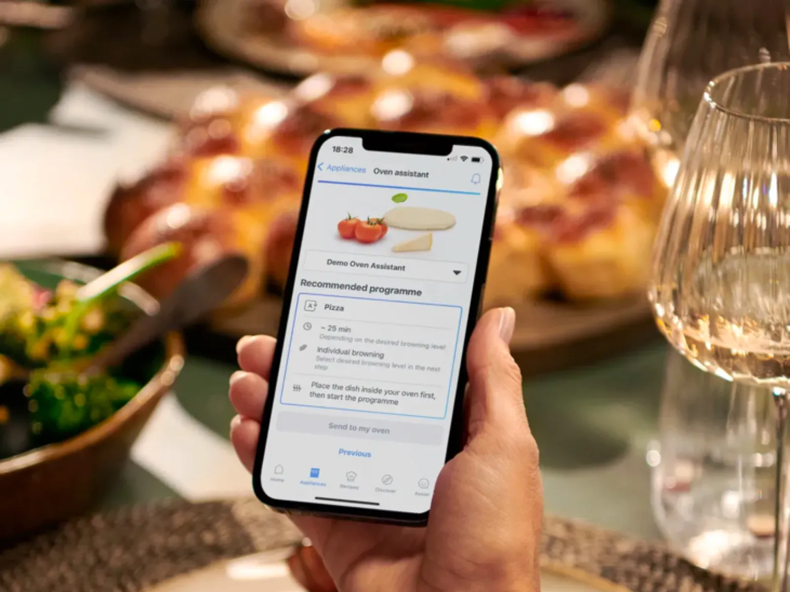 Hand holding a smartphone displaying an oven assistant app with a pizza program selected, blurred dining table in background.