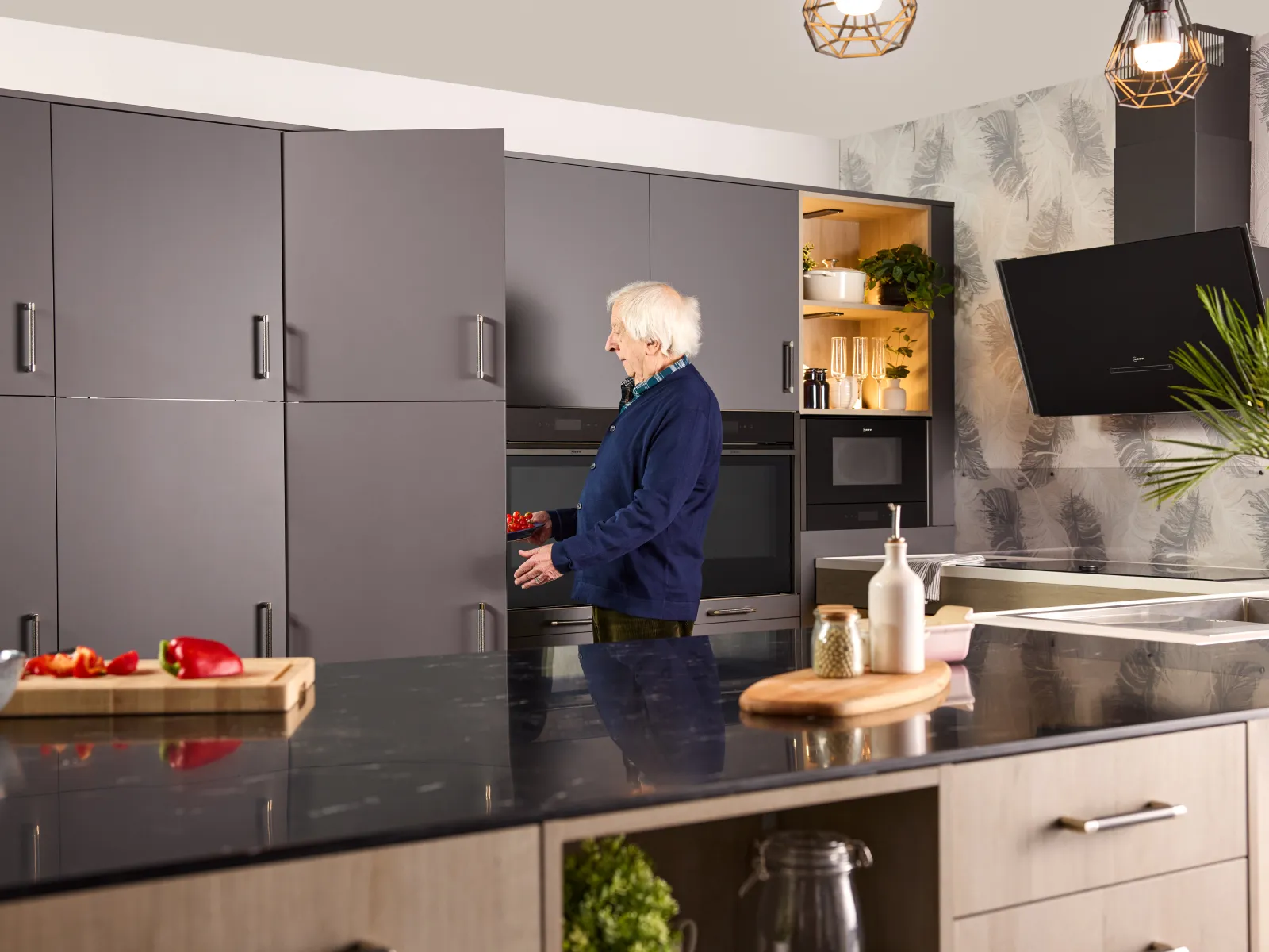 An elderly gentleman standing by grey kitchen cabinets, opening a built-in fridge door and built‑in ovens holding a bowl of cherry tomatoes.