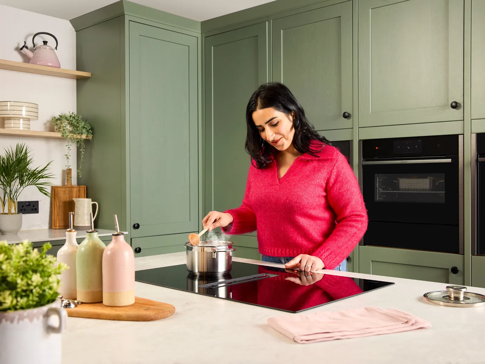 In a green kitchen, a woman with low vision stands at an induction hob. She uses tactile markers on touch controls for safe, precise control.