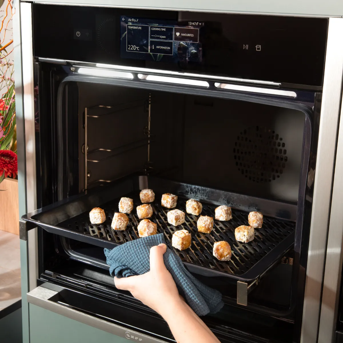 Oven with baked goods, hand reaching in, flowers in vase on counter.