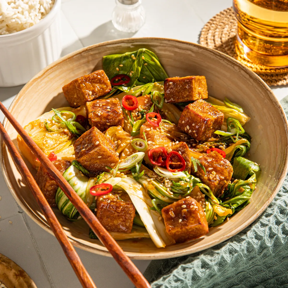 Wooden plate with tofu, vegetables, and chopsticks; glass of amber liquid and bowl of grains in background.