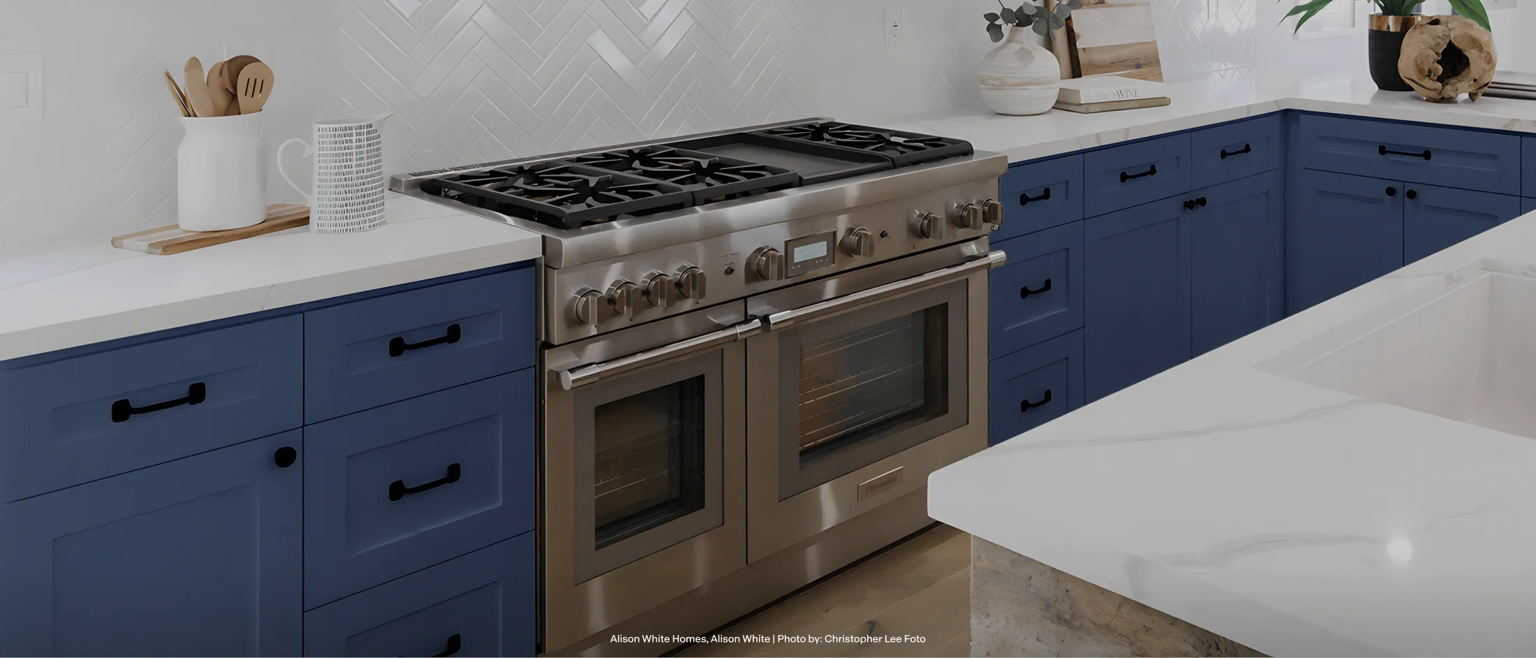 Modern kitchen with blue cabinets, stainless steel dual ovens and stove, white countertops, and various kitchen utensils and decor.