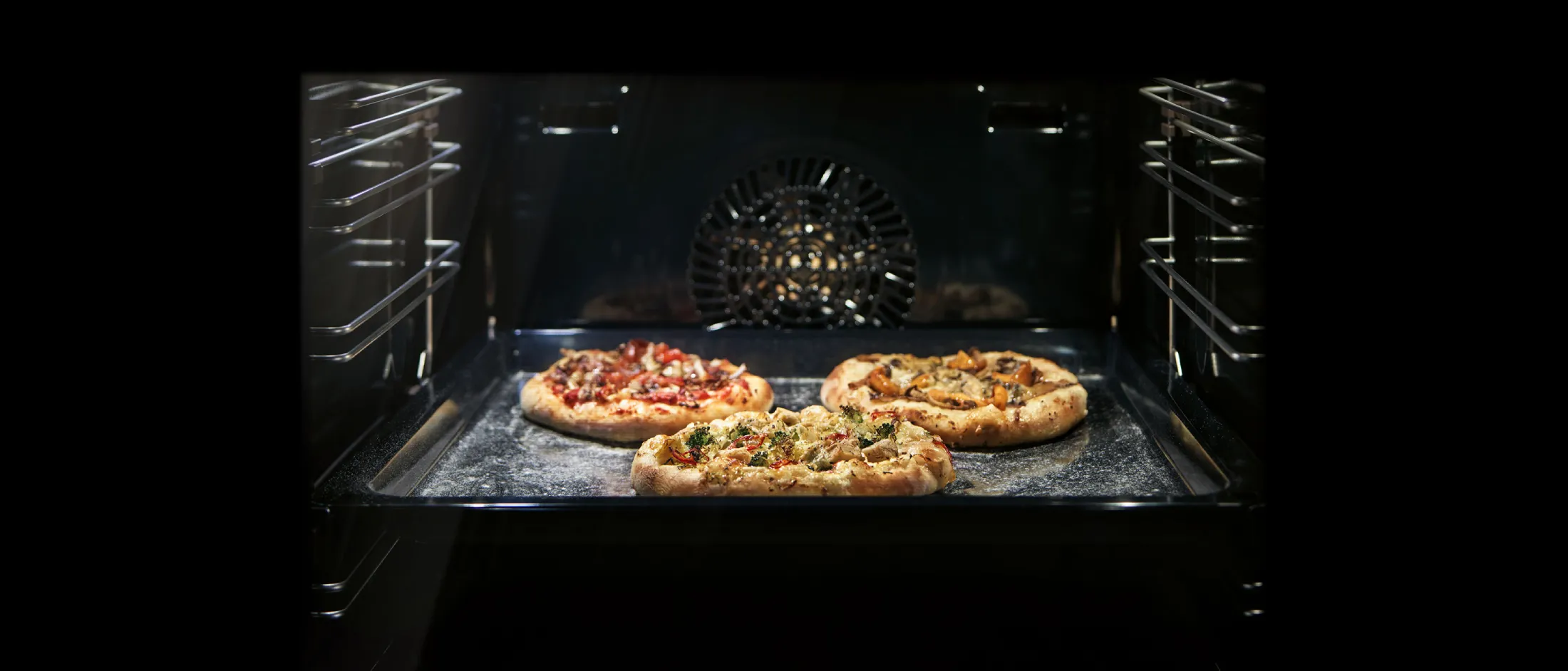 Trois pizzas différentes cuisent simultanément sur une plaque à l’intérieur d’un four, avec le ventilateur de chaleur tournante bien visible.
