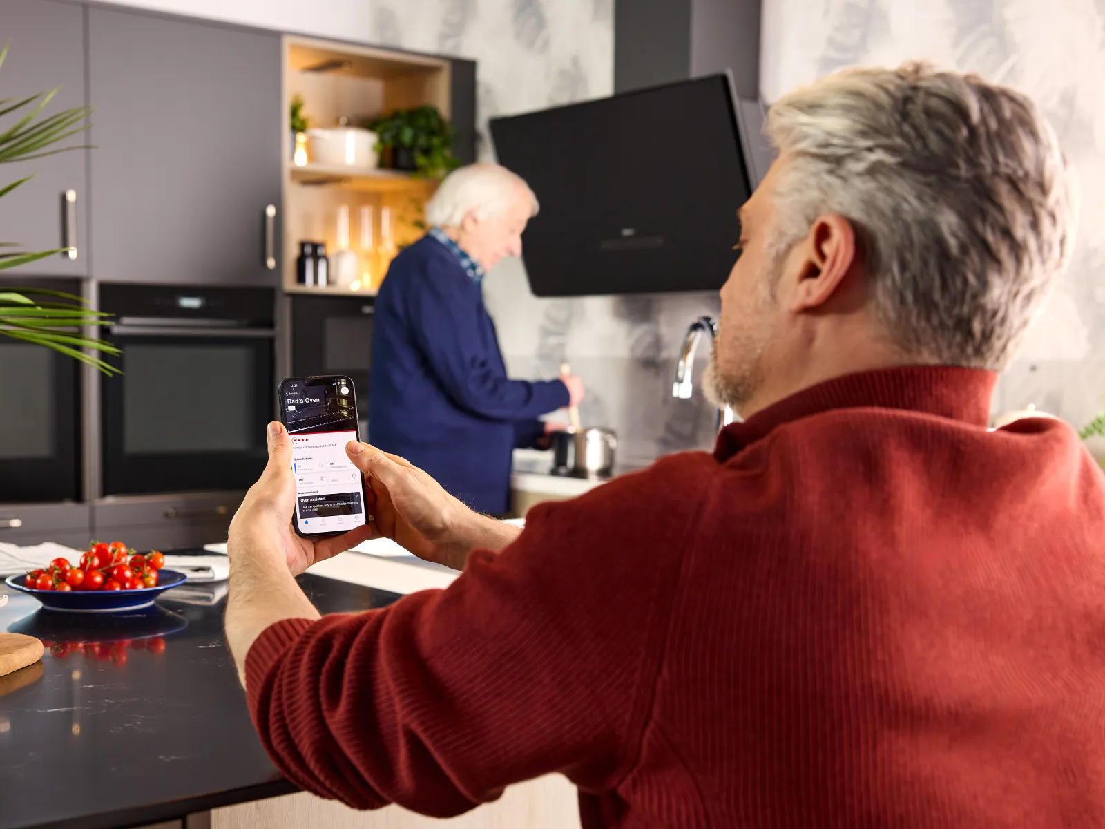 An elderly person cooks at the hob. His son monitors the Home Connect app remotely for added safety and peace of mind.