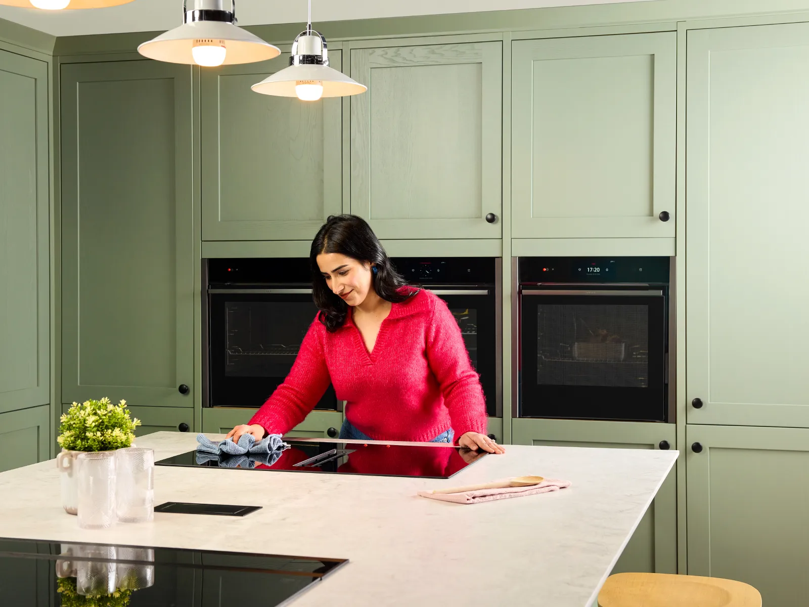 A woman who is visually impaired, wearing a bright pink sweater wiping clean the surface on a black venting induction hob, set into a white kitchen island worktop.