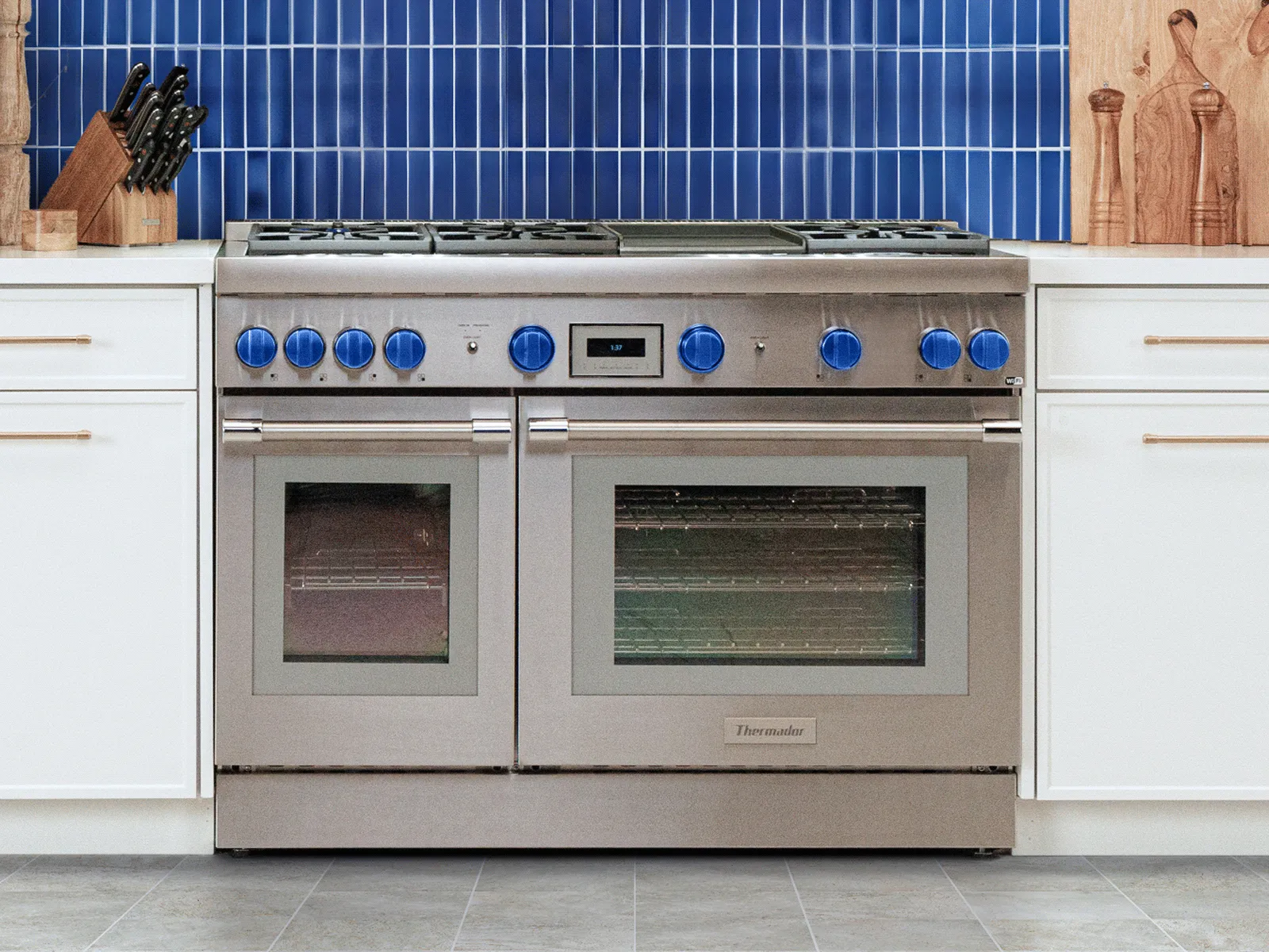Stainless-steel professional double-oven range with six burners and blue control knobs, set between white cabinets against a blue-tiled backsplash.