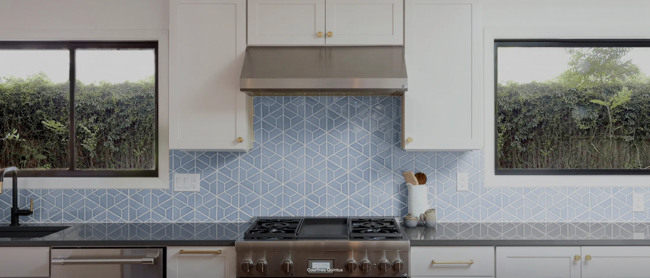 Modern kitchen with stainless steel stove, geometric blue tiled backsplash, white cabinets, black countertop, and windows overlooking greenery