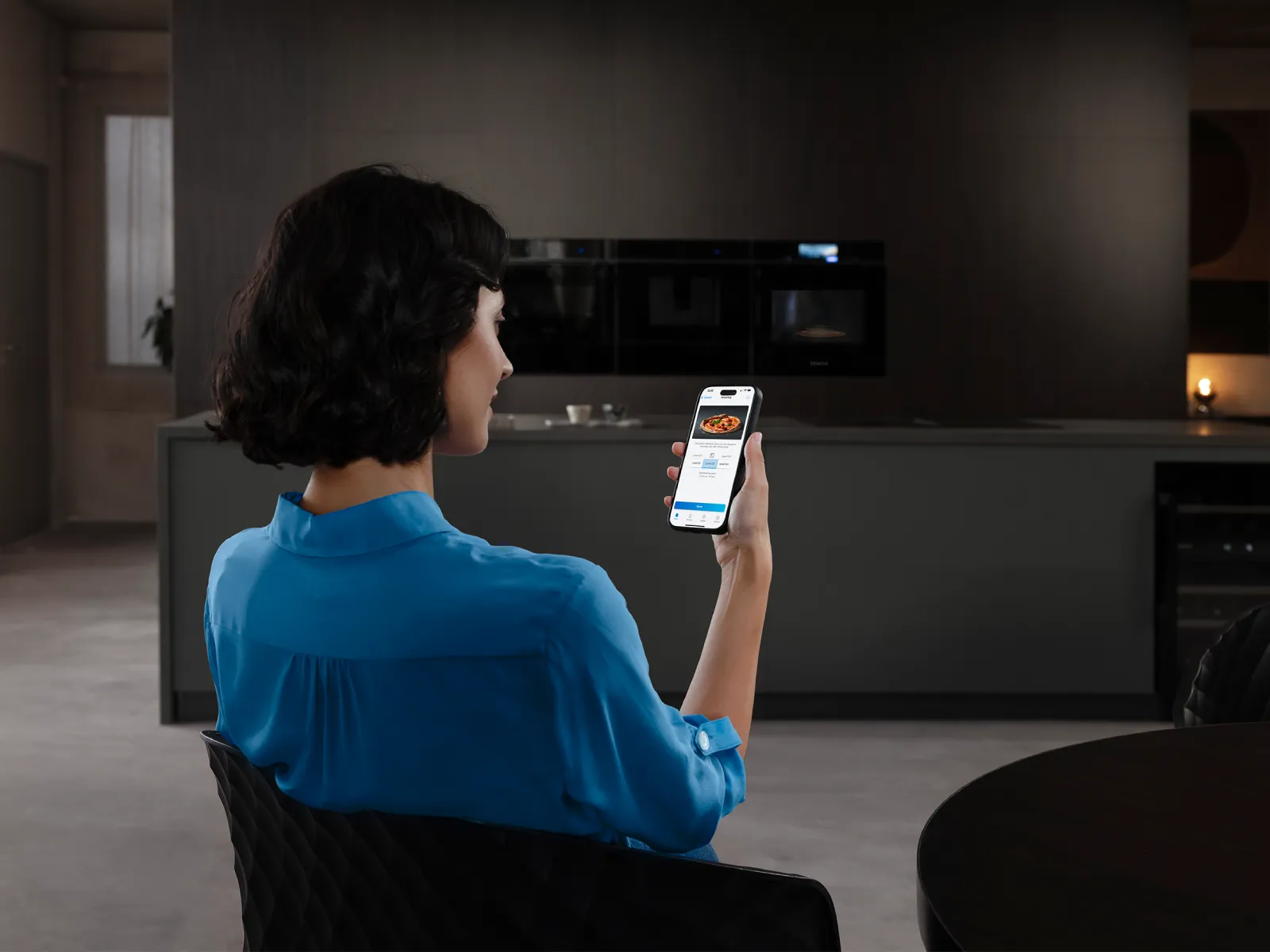 Una persona vestita di blu ha in mano uno smartphone che mostra la schermata di un‘app di food delivery ed è seduto in una cucina arredata in stile moderno.