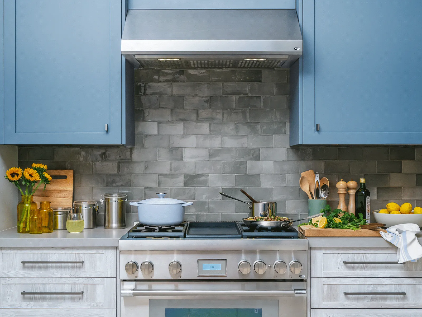 Modern kitchen stove with pots, utensils, sunflowers, fresh herbs, lemons, and blue cabinets against a gray tiled backsplash.