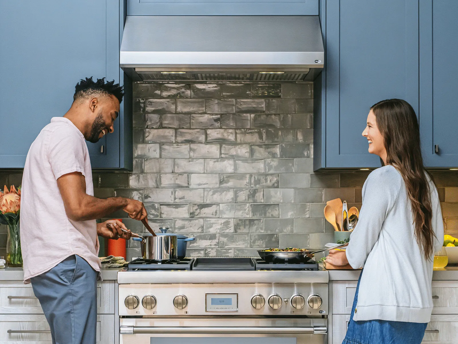 Couple cooking together in a modern kitchen with blue cabinets and a stainless steel stove and hood.
