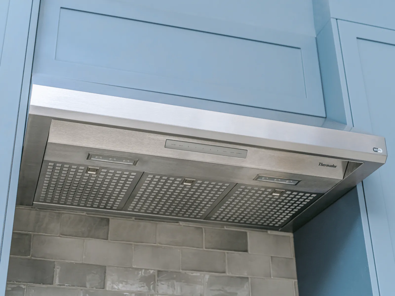 Stainless steel Thermador range hood with built-in lights installed under blue kitchen cabinets and grey tiled backsplash.