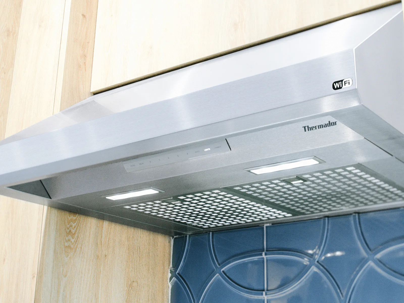 Stainless steel Thermador kitchen range hood with built-in lights, WiFi logo, and modern blue tiled backsplash under wooden cabinets
