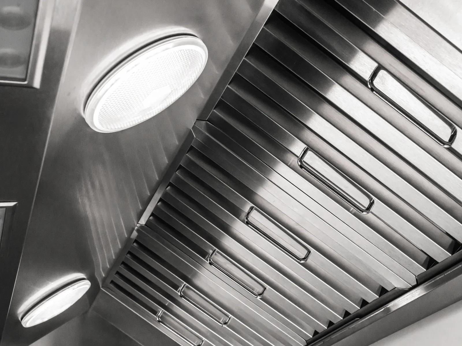 Close-up of a stainless steel commercial kitchen range hood with two round built-in lights and vent slats.