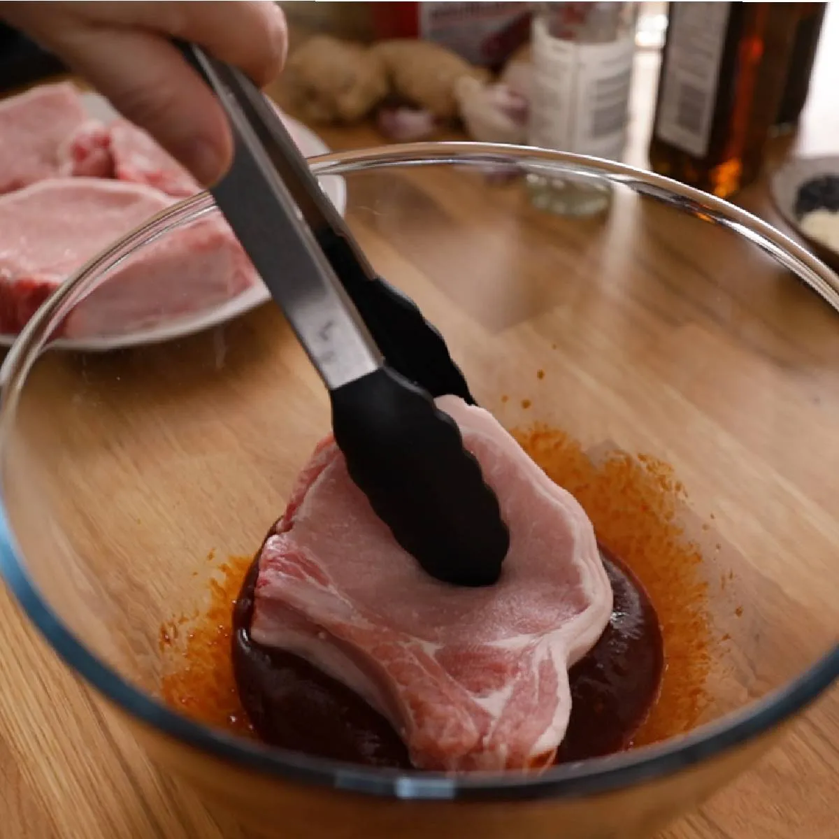 A hand uses black tongs to place a raw pork chop into a clear glass bowl containing a red marinade.