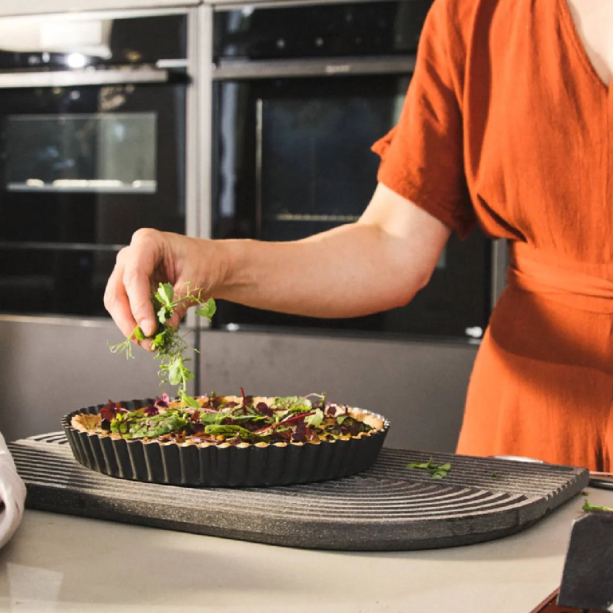 Hands garnishing a savory quiche or tart with fresh greens on a striped platter.