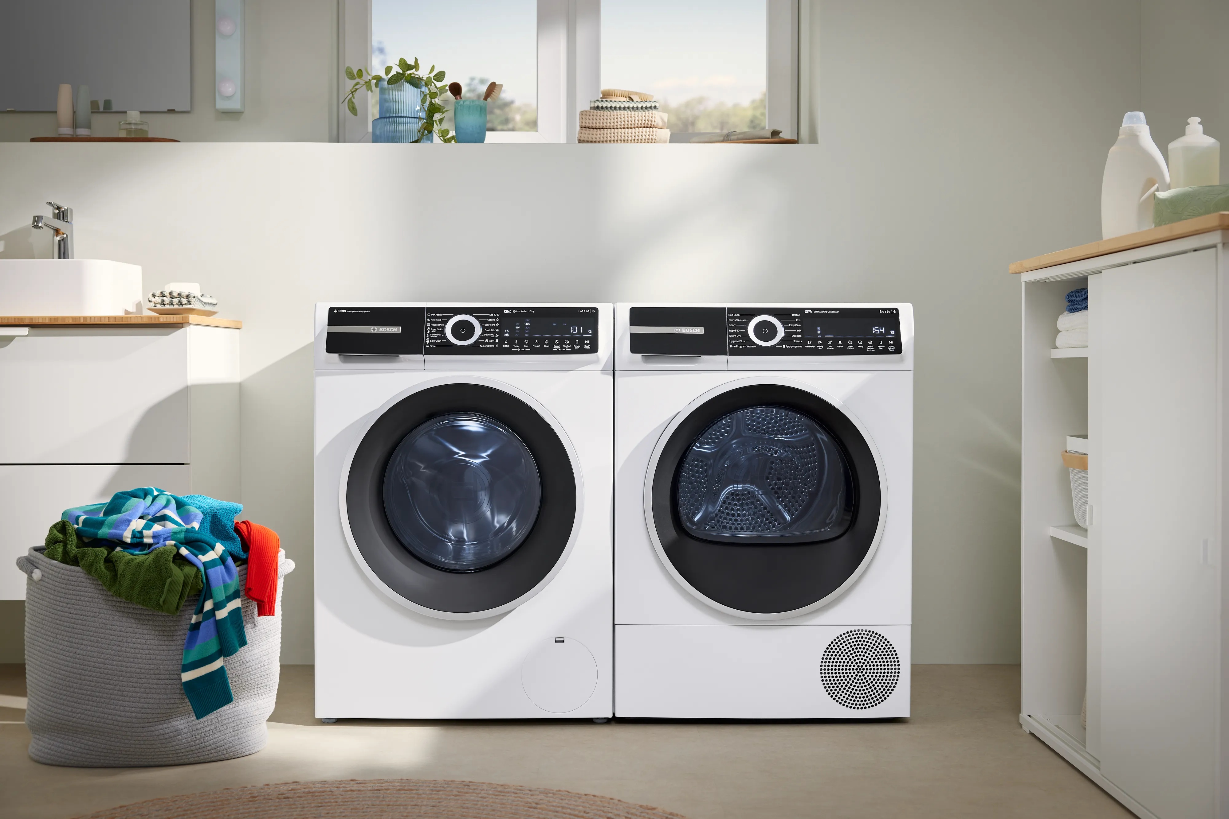 Witte wasmachine en droger naast elkaar in een wasruimte met een wasmand gevuld met kleurrijke kleding links