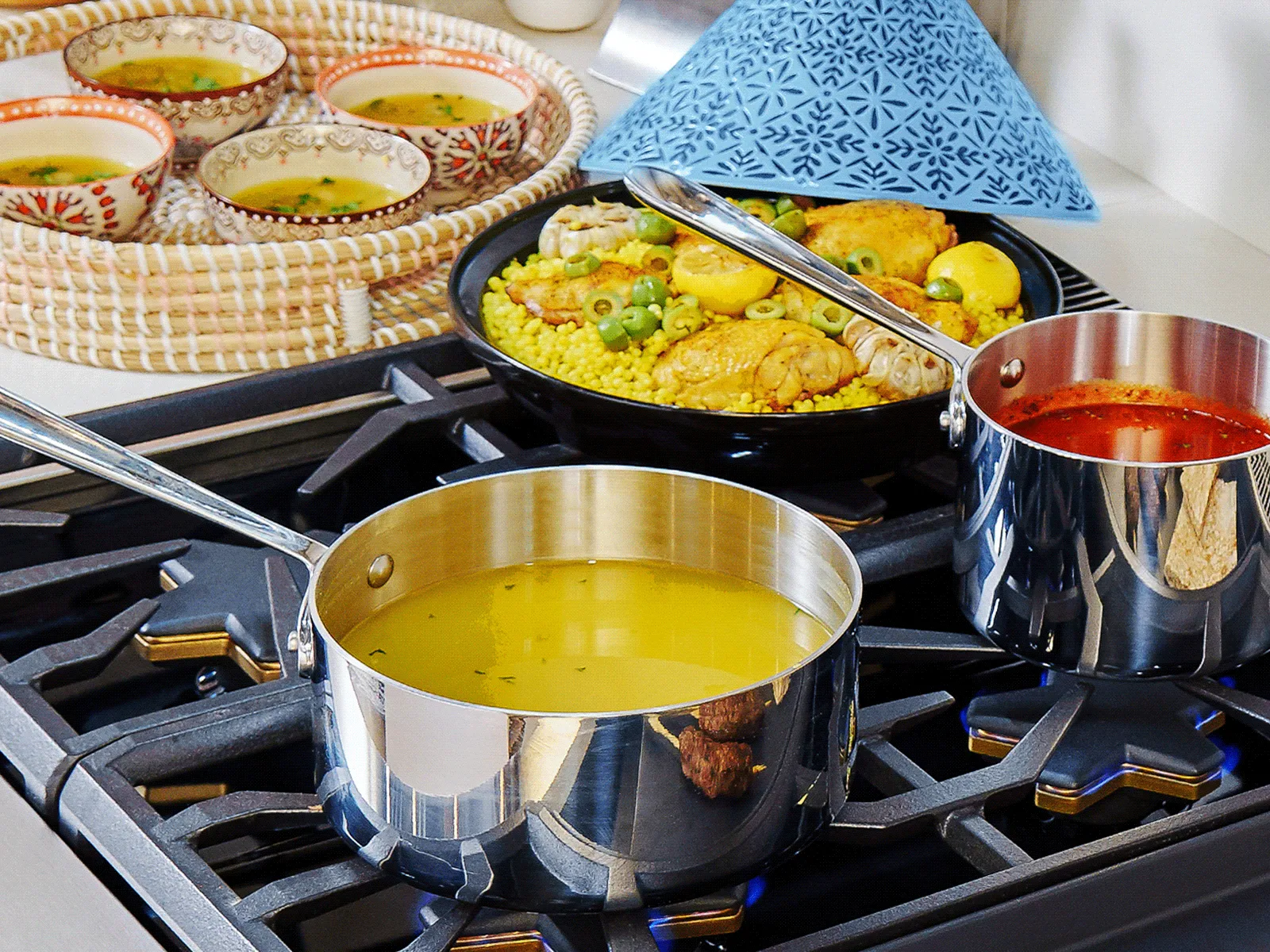 Stovetop scene with pots of yellow lentil soup and red sauce, a vegetable dish under a blue cover, and small bowls of spices nearby.