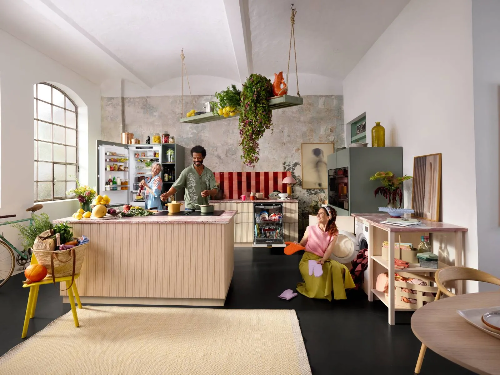 Spacious, sunlit kitchen with a large arched window: a man cooks at a central island while a Lady opens the fridge and pours a drink, and another Lady is unloading the washing machine holding bright orange oven mitts; hanging plants, bowls of fruit, and an open dishwasher complete the scene.