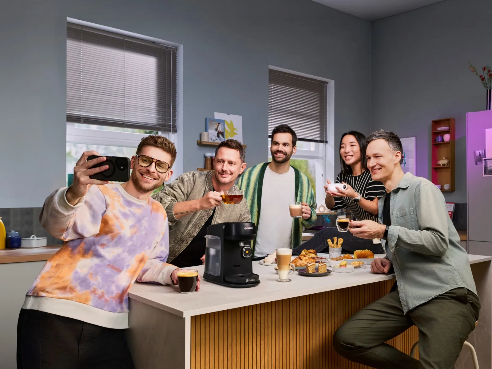 Friends gathered at a kitchen counter with a Bosch TASSIMO Harmony coffee machine, holding freshly made hot beverages and sharing snacks