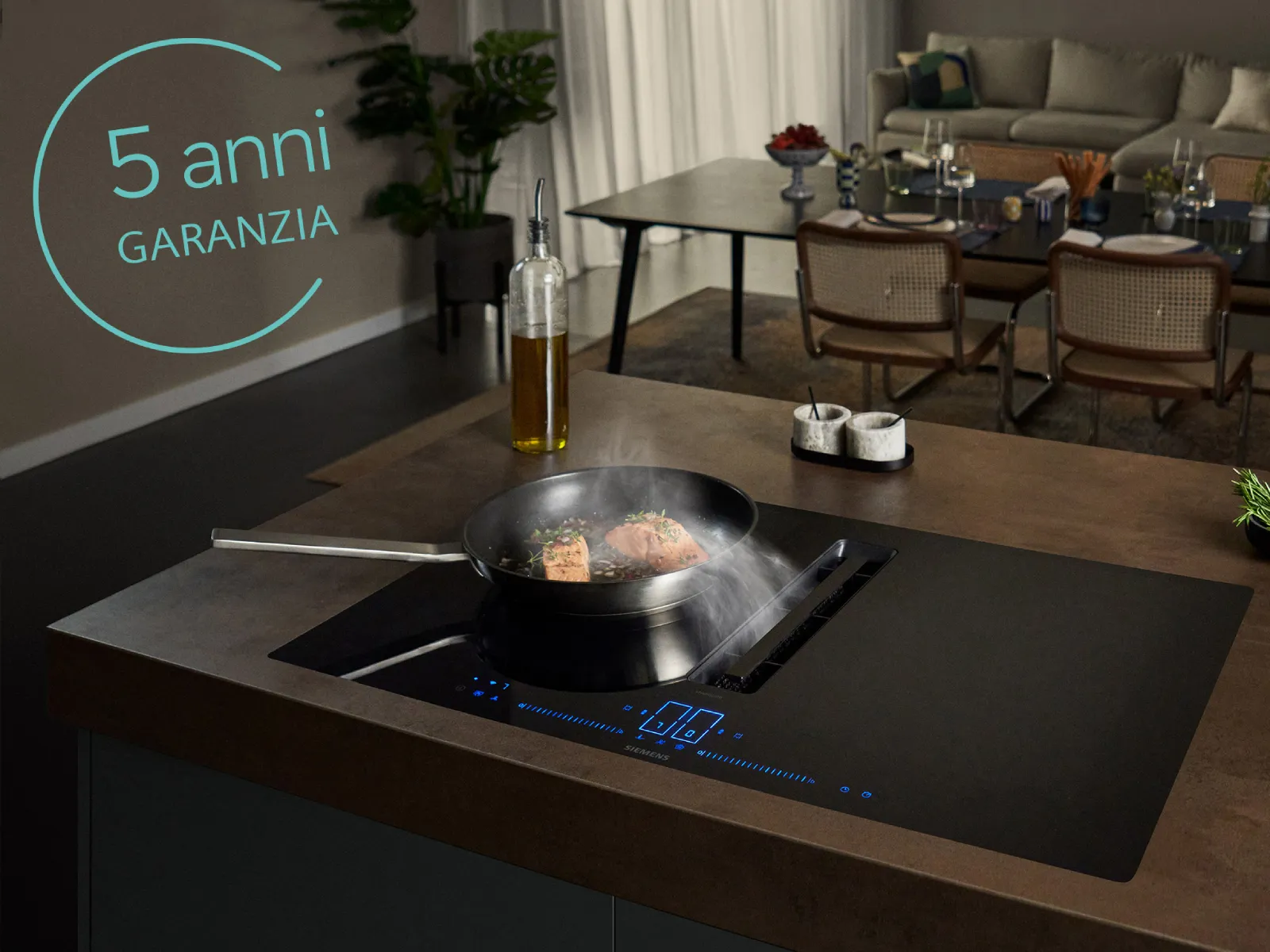 Cucina moderna con padella affumicata su piano cottura a induzione, tavolo da pranzo sullo sfondo e sovrapposizione di testo "5 anni garanzia".