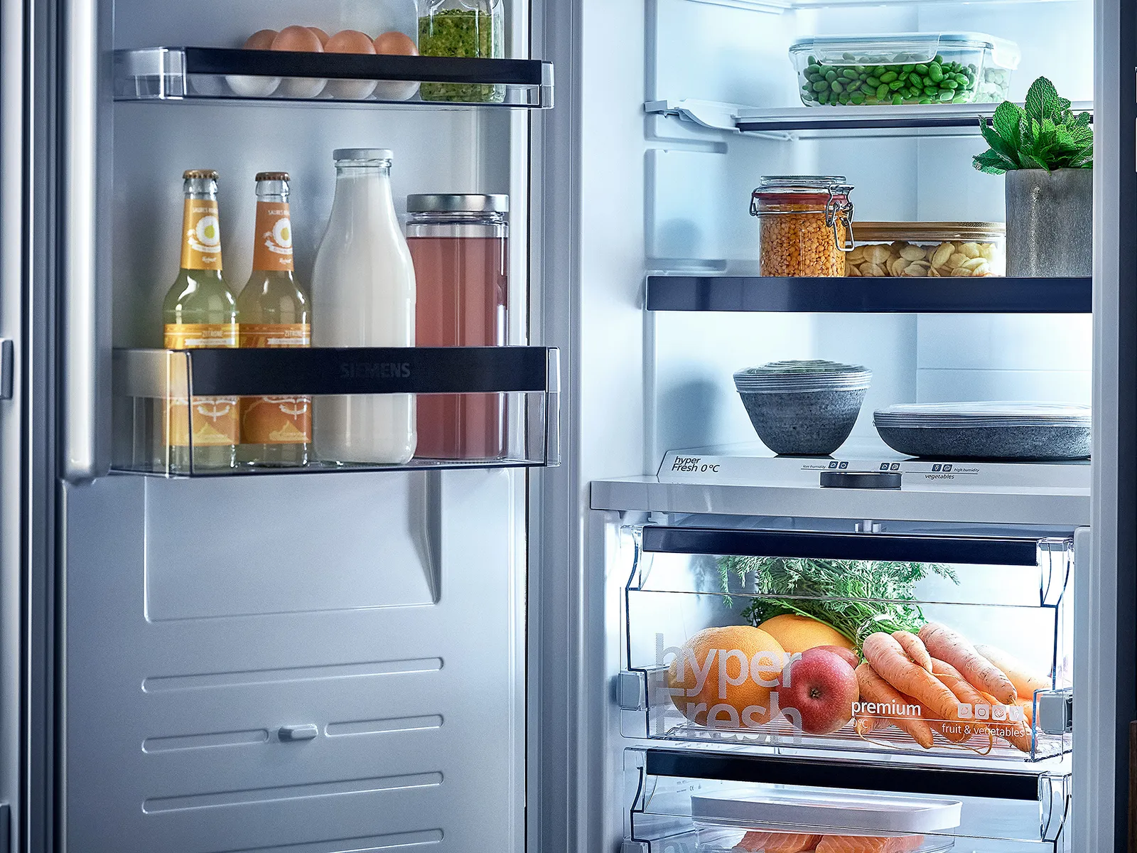 Ein hell erleuchteter, offener Kühlschrank mit perfekt organisierten Lebensmitteln und frischen Zutaten, direkt neben einem perfekt integrierten Siemens Einbackofen.