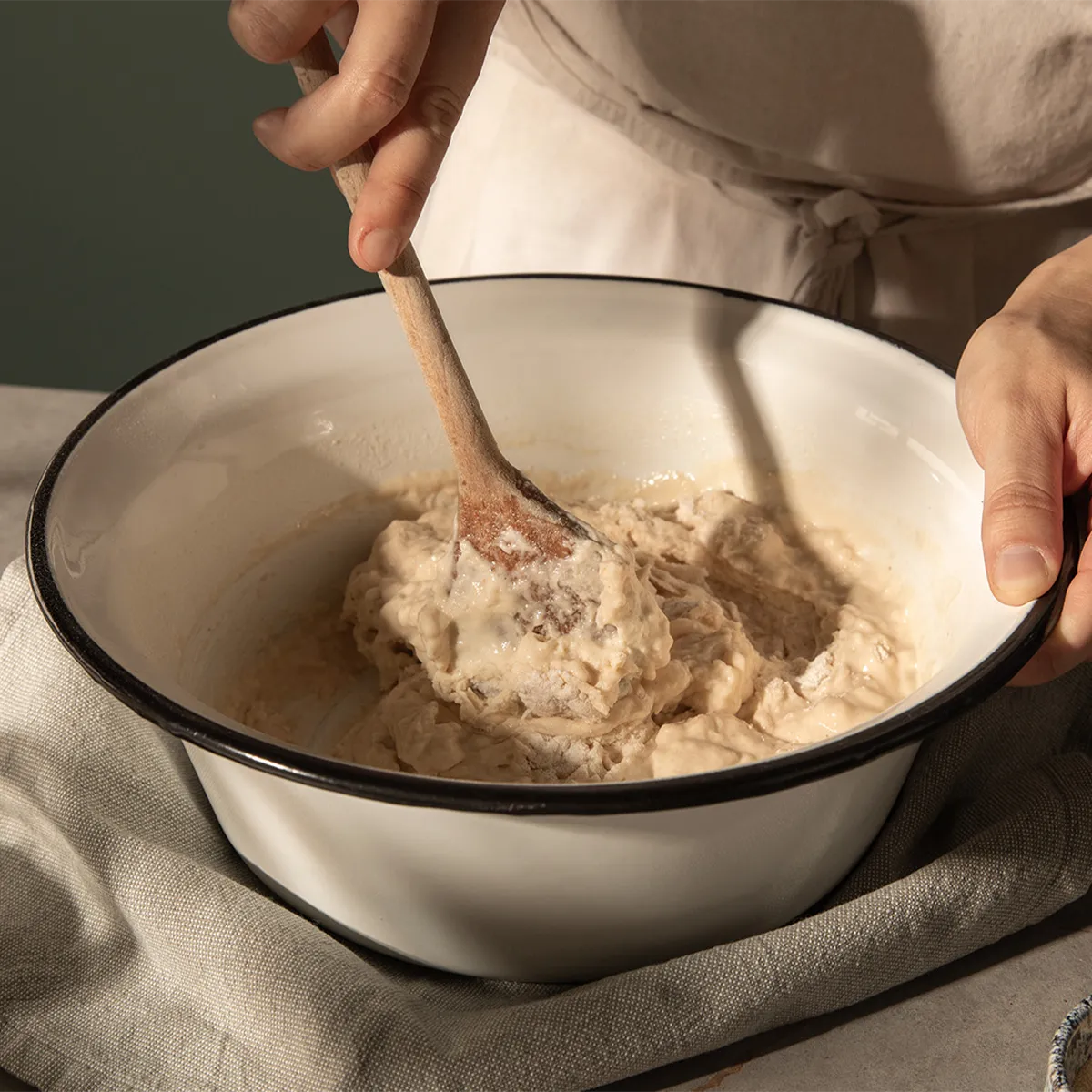 Hands stirring thick dough in a white bowl with a wooden spoon, set on a cloth-covered surface.
