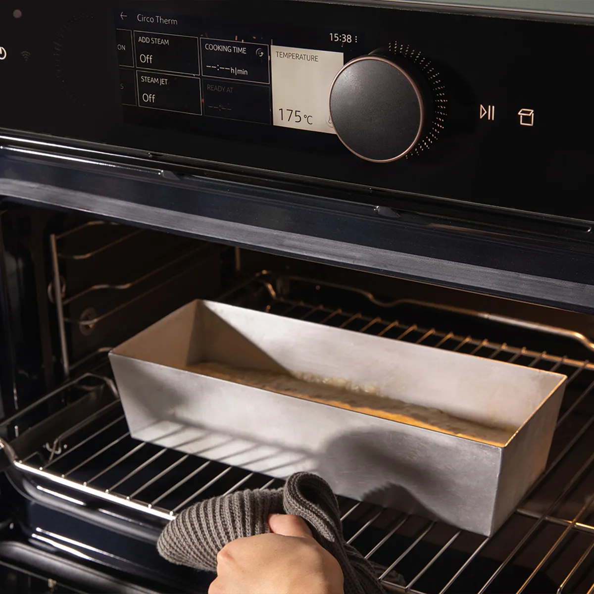 Hand placing a metal loaf pan with batter into an oven set at 175°C using a gray oven mitt.