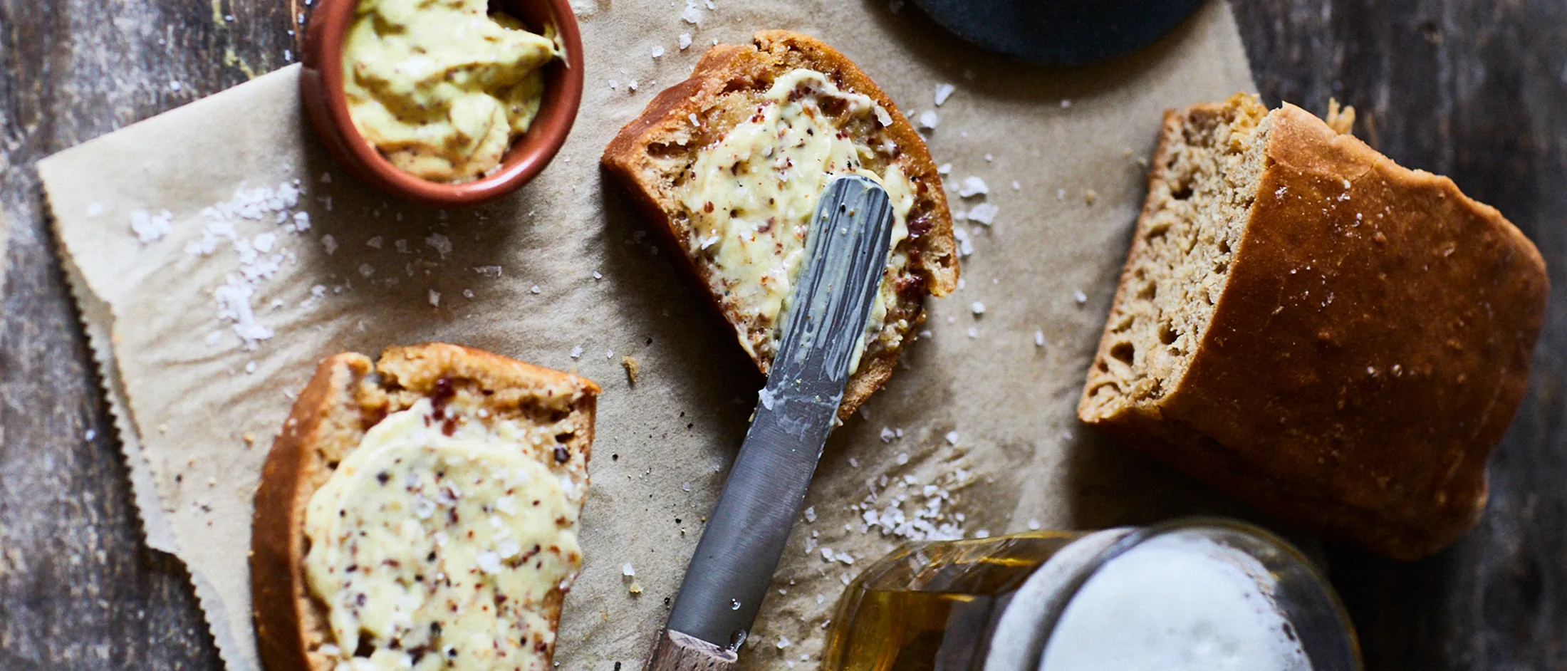 Freshly baked bread slices spread with herb butter on parchment, accompanied by a knife and a drink on a rustic wooden table.