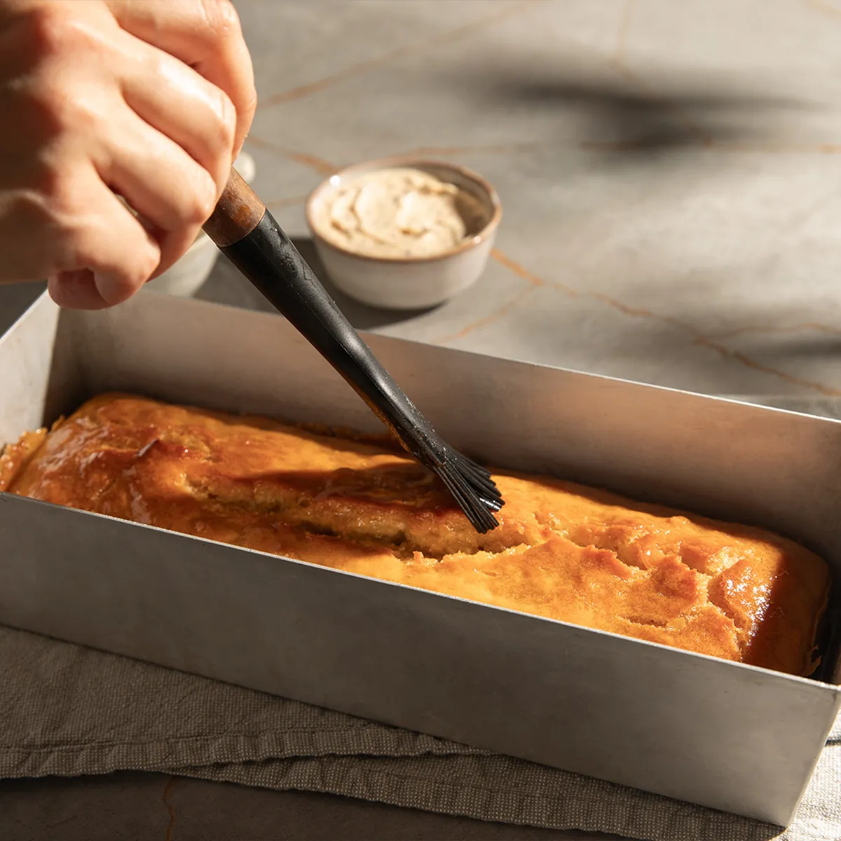 Hand brushing glaze onto freshly baked golden loaf cake in a metal pan on a gray cloth and marble surface.