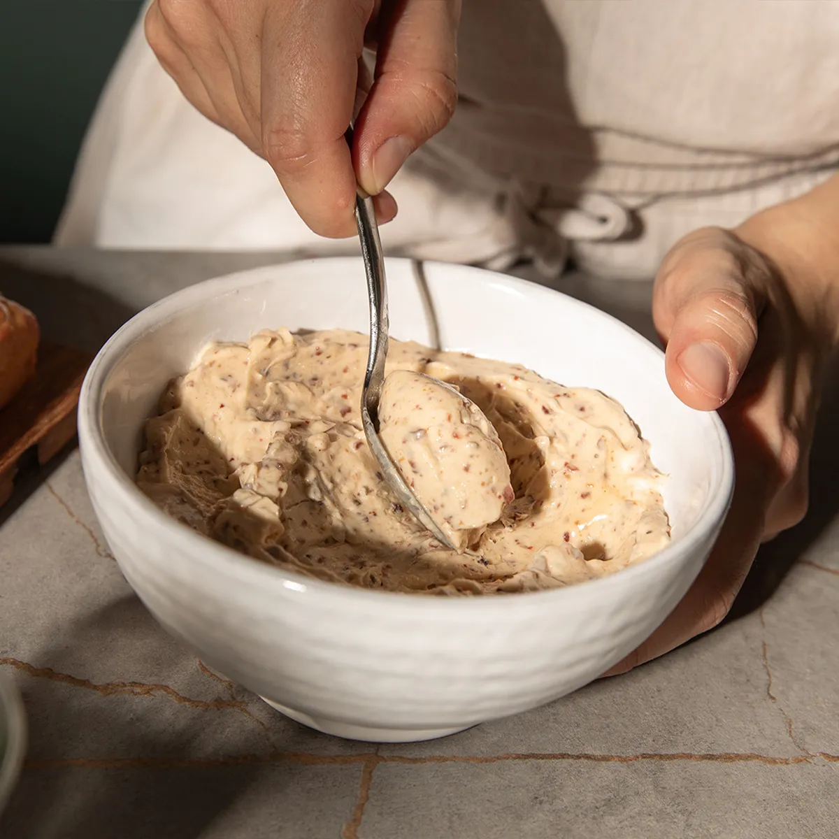 Hand holding a spoon scooping creamy, nut-speckled spread from a white bowl on a stone countertop.